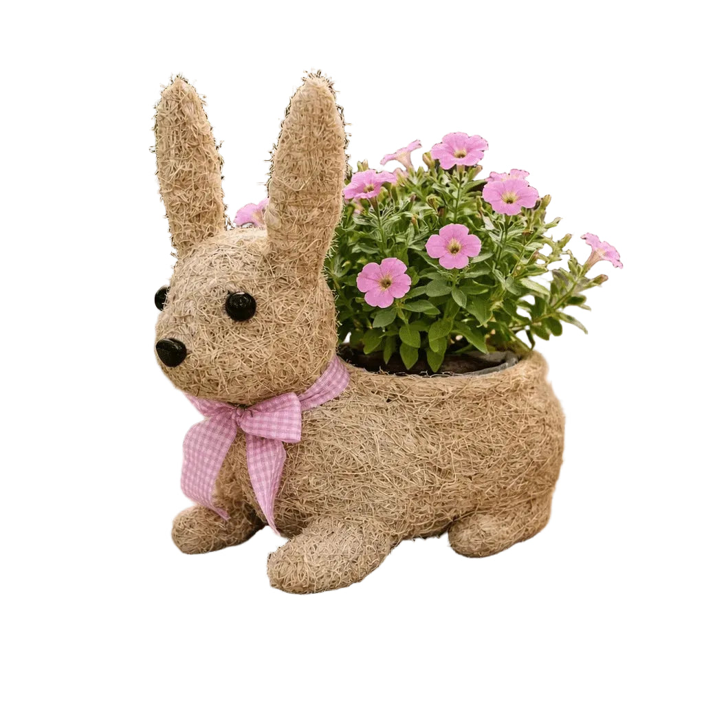 A rustic rabbit-shaped planter made from natural coir rope, decorated with a pink gingham ribbon bow, holding a vibrant green plant with pink flowers, set on a weathered wooden table in an outdoor garden setting.